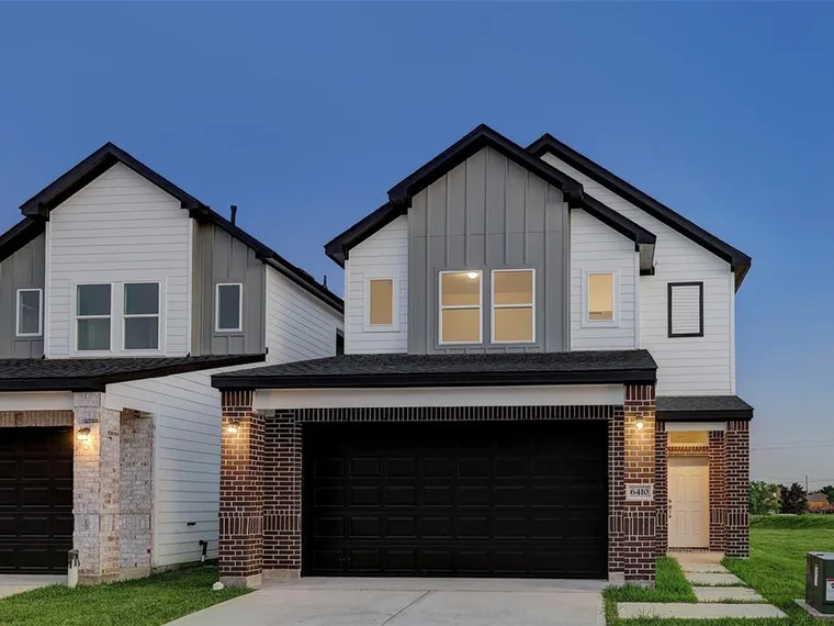 New two-story home with white and gray siding, a brick-framed black garage door and a lit entry at dusk at 6410 Leopold Star Ln, Houston, Texas